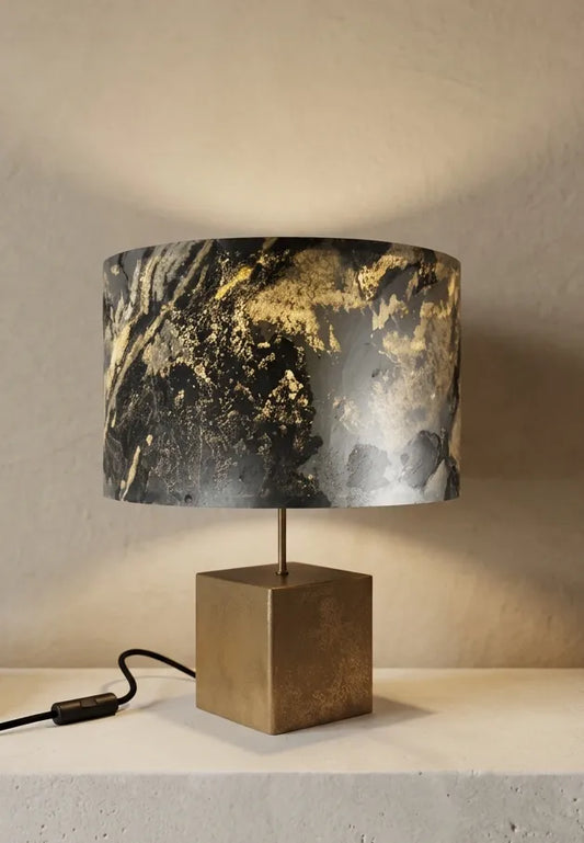 Lampe en acier revêtu de poudre de bronze et son abat-jour en feuille d'ardoise rustique allumée, modèle Cassini posée sur une console en pierre blanche contre un mur de chaux beige