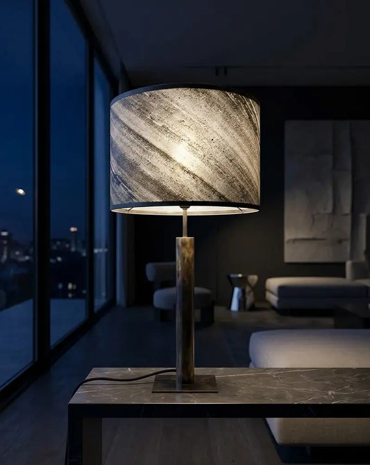 Mise en scène de la lampe lampe en acier patiné abat-jour en ardoise zébrée artisanale sur une console en pierre noire, modèle Rosalynd par l'atelier Maus dans un salon au design contemporain la nuit. scène virtuelle, produit réel