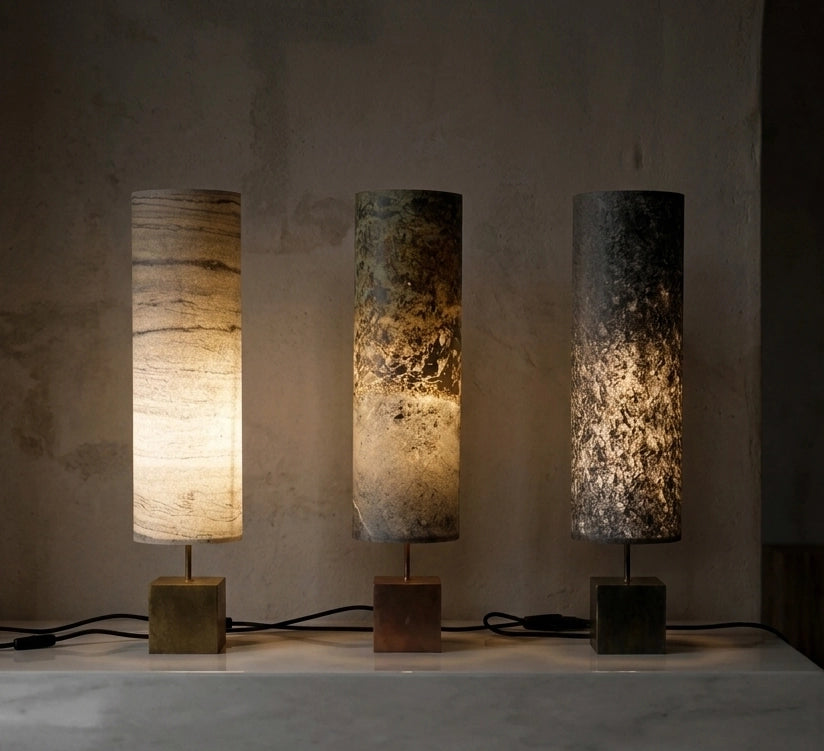 3 lampes en acier patiné  et feuille de pierre  - marbre, ardoise et mica - Halley Nova de l'atelier Maus allumées. les lampes sont posées sur une console en pierre polie de couleur claire contre un mur en béton brut.