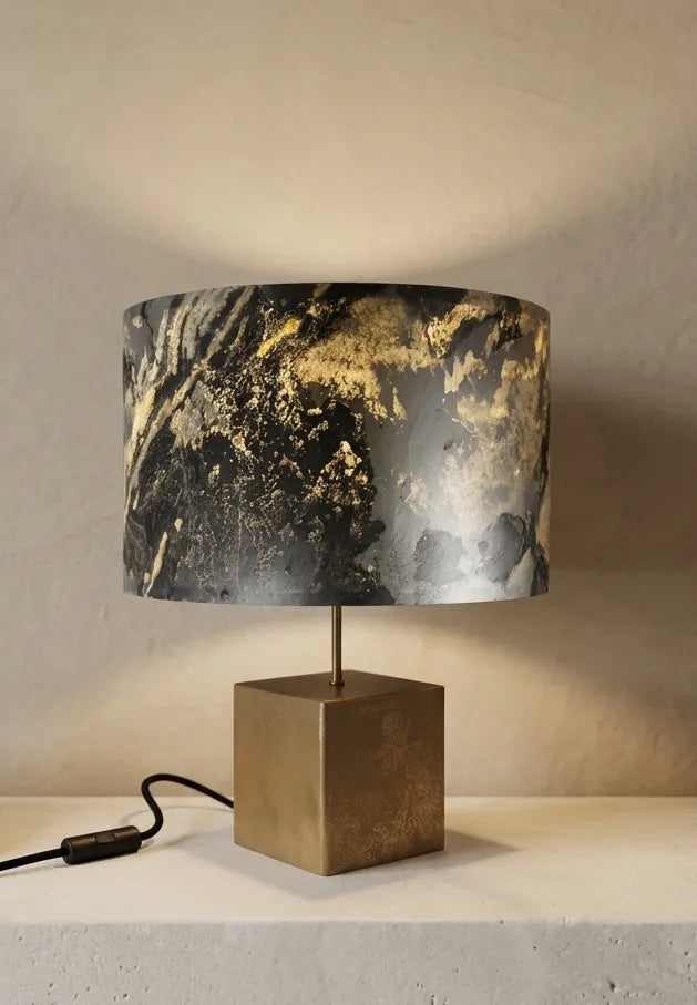 Lampe en acier revêtu de poudre de bronze et son abat-jour en feuille d'ardoise rustique allumée, modèle Cassini posée sur une console en pierre blanche contre un mur de chaux beige