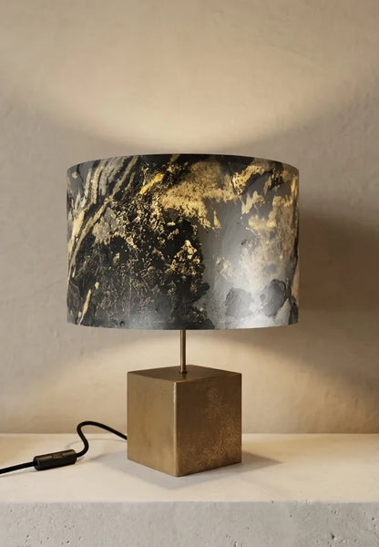 Lampe en acier revêtu de poudre de bronze et son abat-jour en feuille d'ardoise rustique allumée, modèle Cassini posée sur une console en pierre blanche contre un mur de chaux beige