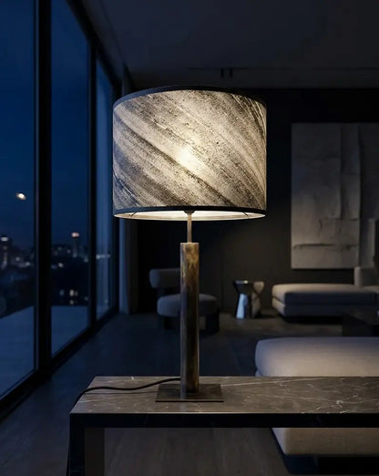 Mise en scène de la lampe lampe en acier patiné abat-jour en ardoise zébrée artisanale sur une console en pierre noire, modèle Rosalynd par l'atelier Maus dans un salon au design contemporain la nuit. scène virtuelle, produit réel